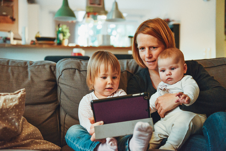 Mum sitting on sofa with baby on lap and toddler next to her looking at a tablet