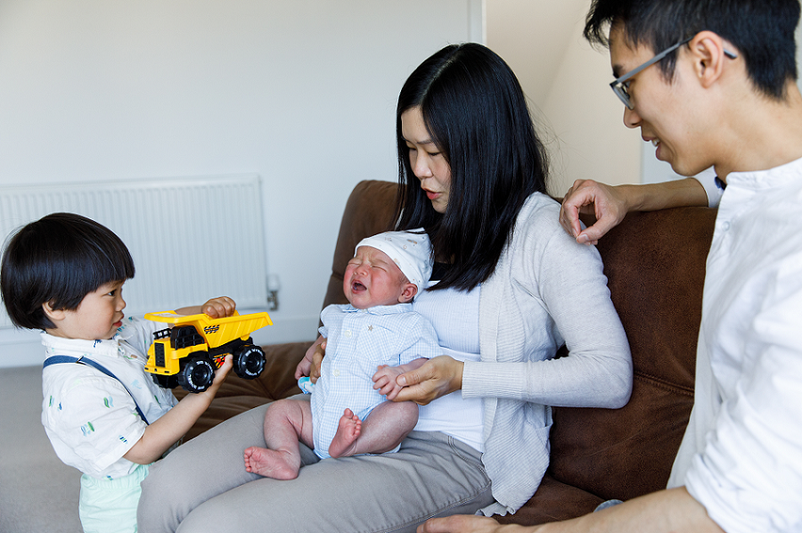 Mum holding crying baby sitting next to dad, with toddler holding toy truck